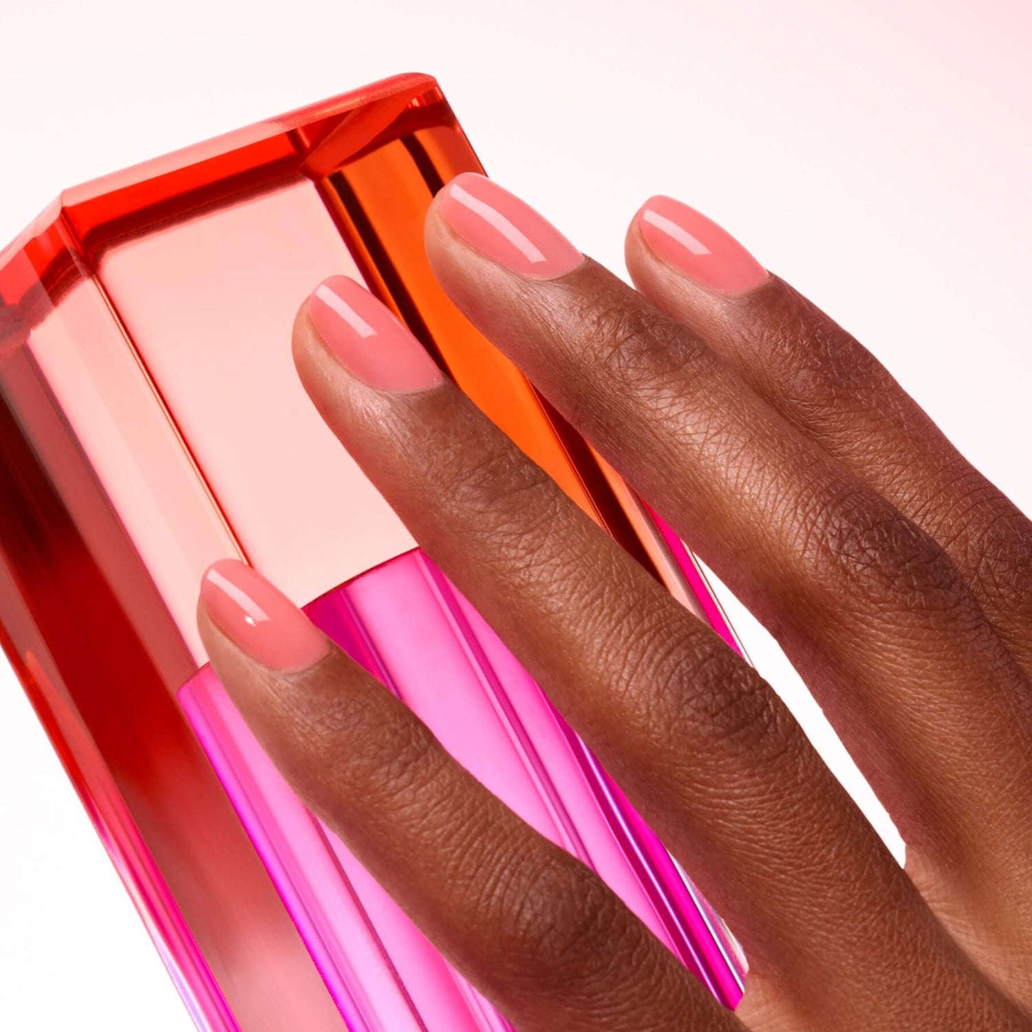 A hand with neatly manicured, glossy Sugar Plum nail polish by Manucurist in pink rests on a vibrant pink and orange geometric bottle against a light background.