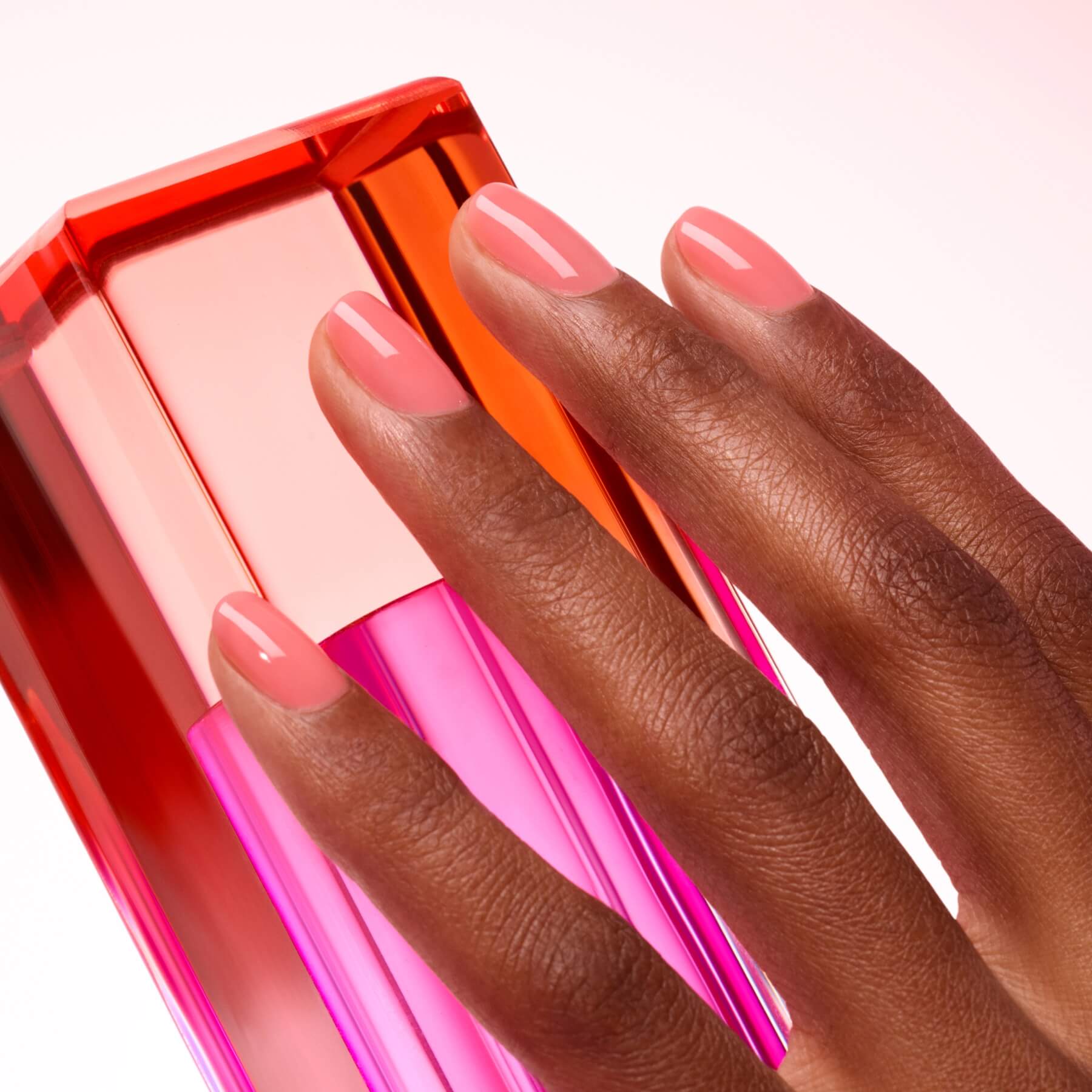 A hand with neatly manicured, glossy Sugar Plum nail polish by Manucurist in pink rests on a vibrant pink and orange geometric bottle against a light background.