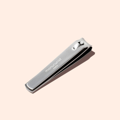 A silver Manucurist Nail Clipper, featuring the brand name engraved on its surface, is placed on a light beige background.