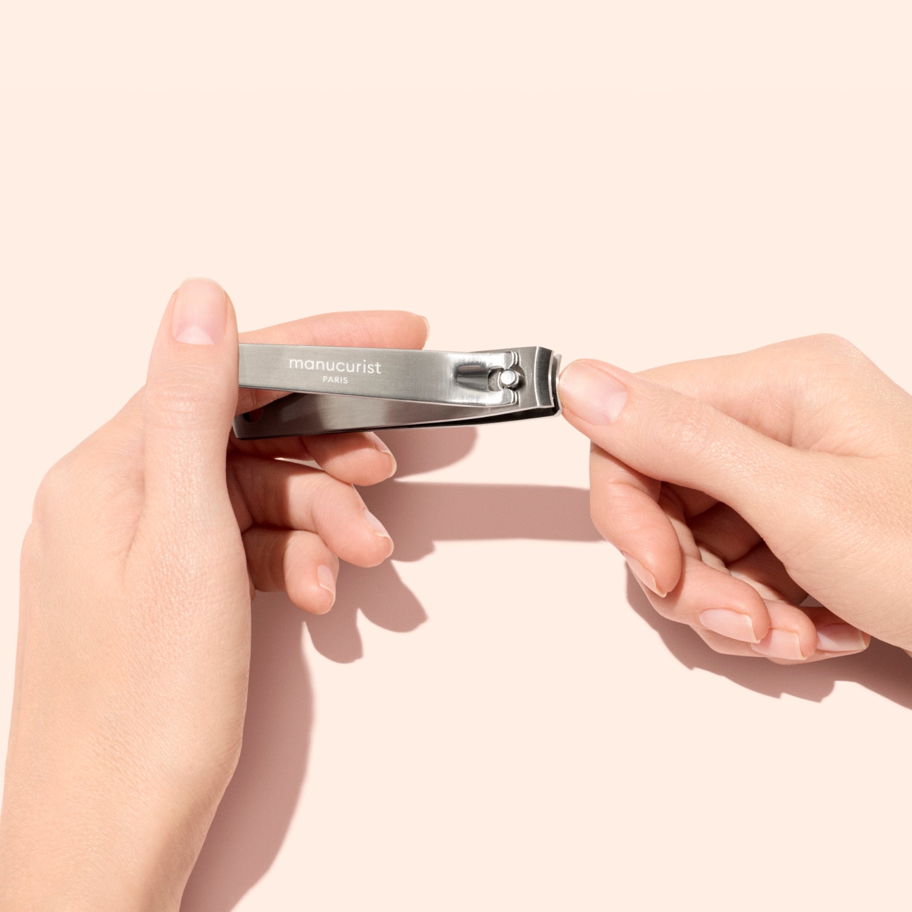 A person holds a Manucurist Nail Clipper in one hand, positioning it to trim the nails of the other hand against a pale pink background.