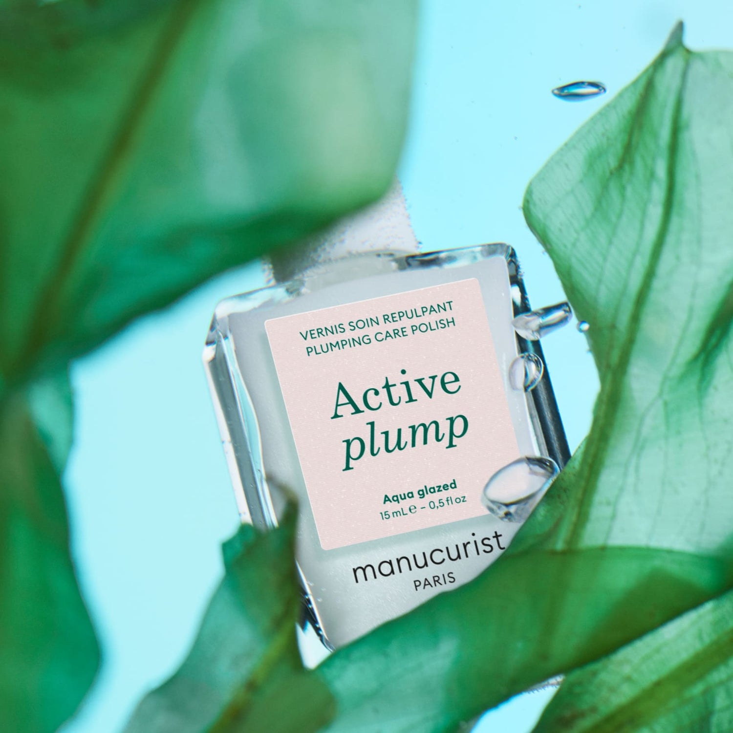 A bottle of Manucurist Active Plump Aqua Glazed nail polish sits among green leaves on a blue background, with water droplets on the bottle.