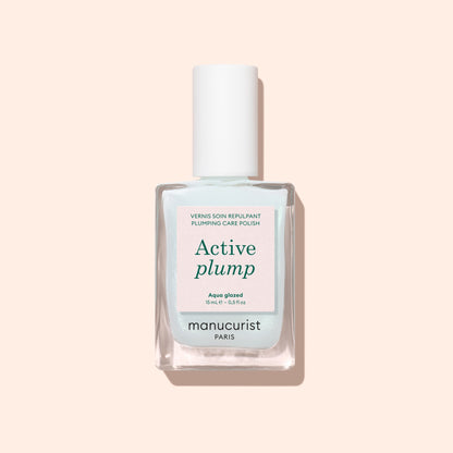 A bottle of Manucurist Active Plump Aqua Glazed nail polish with a white cap sits on a light beige background. The pale blue and white label features green and pink text, highlighting hydration for beautifully healthy nails.