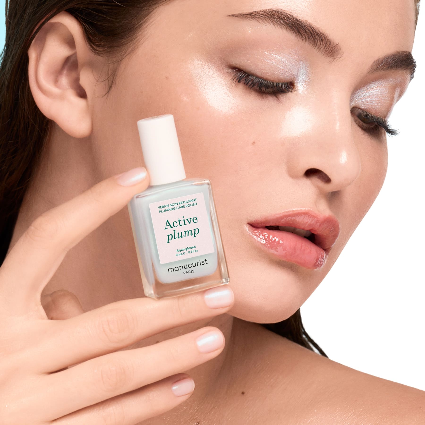 A woman with dewy, glowing skin and shimmery eyeshadow holds up a bottle labeled Active Plump Aqua Glazed by Manucurist near her face, displaying hydrated nails with a pearly finish.