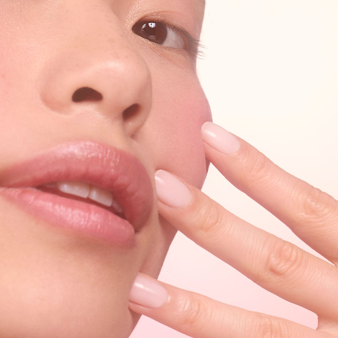 Close-up of a persons face and hand, showing smooth skin, natural makeup, and neatly manicured fingernails painted in a light nude color. The overall lighting is soft and pink-toned.