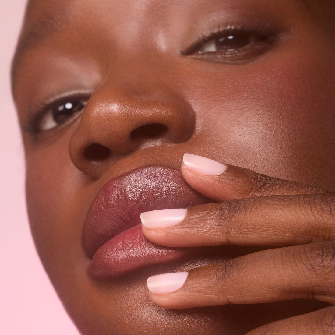 A close-up of a person with smooth skin and glossy lips touches their lips with neatly manicured fingers, highlighting Manucurist’s Active Blur nail-enhancing pale pink polish against a soft pink background.