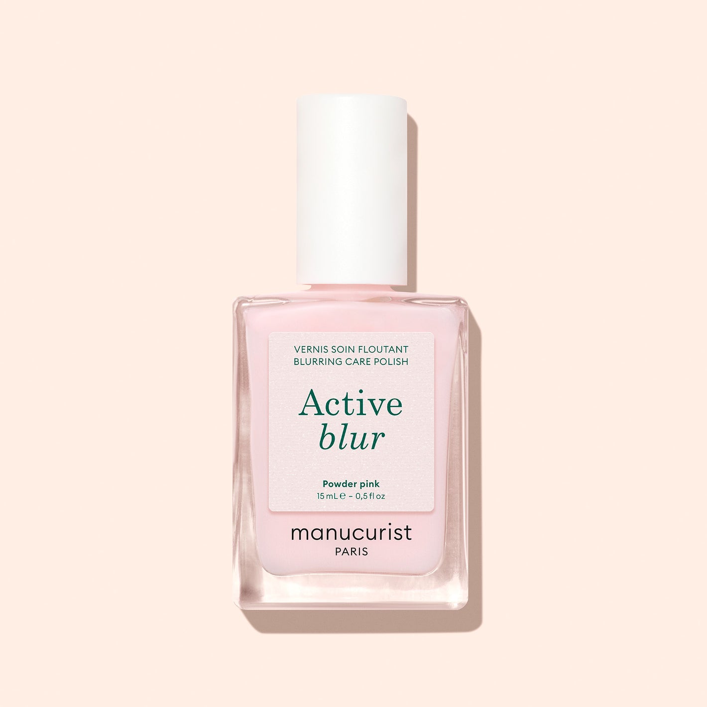 A bottle of Manucurist Active Blur Blurring Care Polish in powder pink, with a white cap and green text on a pastel pink background. The velvety matte formula leaves nails perfectly polished.