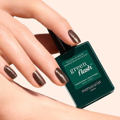 A hand with short, neat nails painted in shimmery brown with gold and silver shimmer holds a dark green bottle of Manucurist Glitter Top Coat against a light background.