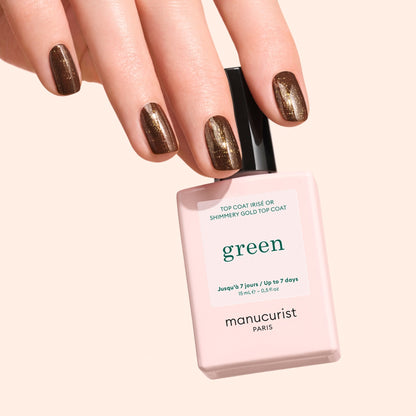 A hand with short, shimmery dark brown nails holds a bottle of Manucurist Glitter Top Coat, featuring a gel-like finish, against a light peach background.