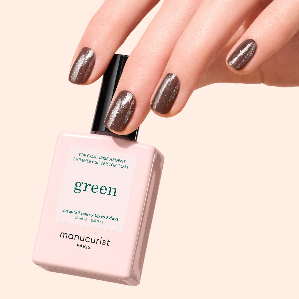 Une main aux ongles manucurés, vernis d’un brun scintillant, tient un flacon de Manucurist Glitter Top Coat, mettant en valeur une finition éclatante effet gel sur un fond beige clair.