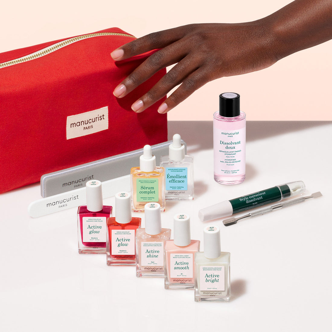 A hand reaches toward a red pouch from the Manucurist Super Ultimate Active Manicure Kit, which is artfully arranged on a white surface and includes all the nail care essentials—cuticle remover, polishes, files, removers, and serums—for healthy nails.