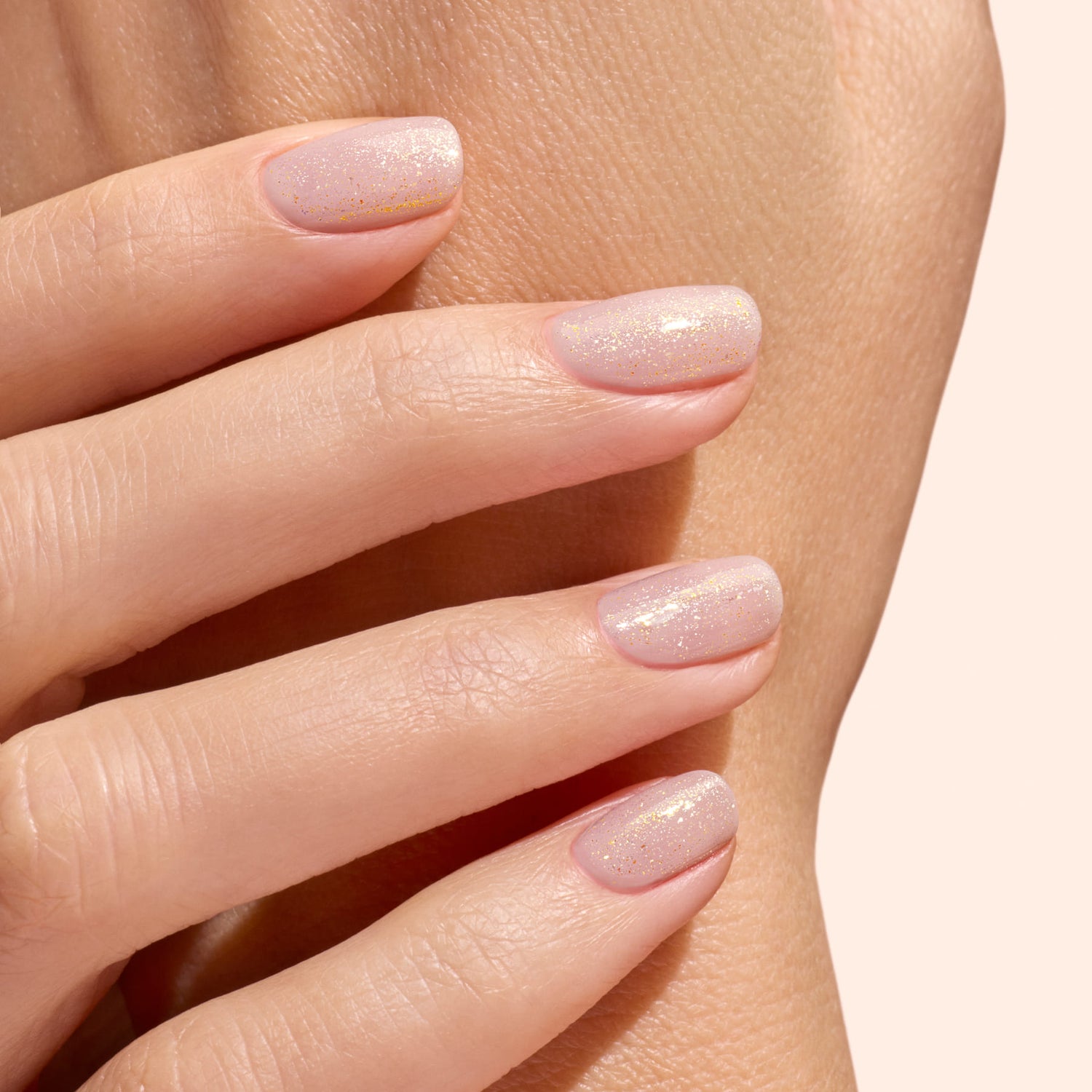 A close-up of neatly manicured nails, finished with Manucurist’s Glitter Top Coat for a gel-like shine and subtle gold shimmer, gently resting on another hand against a soft beige background.