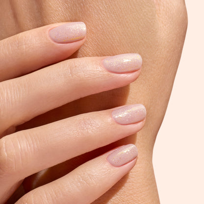 A close-up of neatly manicured nails, finished with Manucurist’s Glitter Top Coat for a gel-like shine and subtle gold shimmer, gently resting on another hand against a soft beige background.