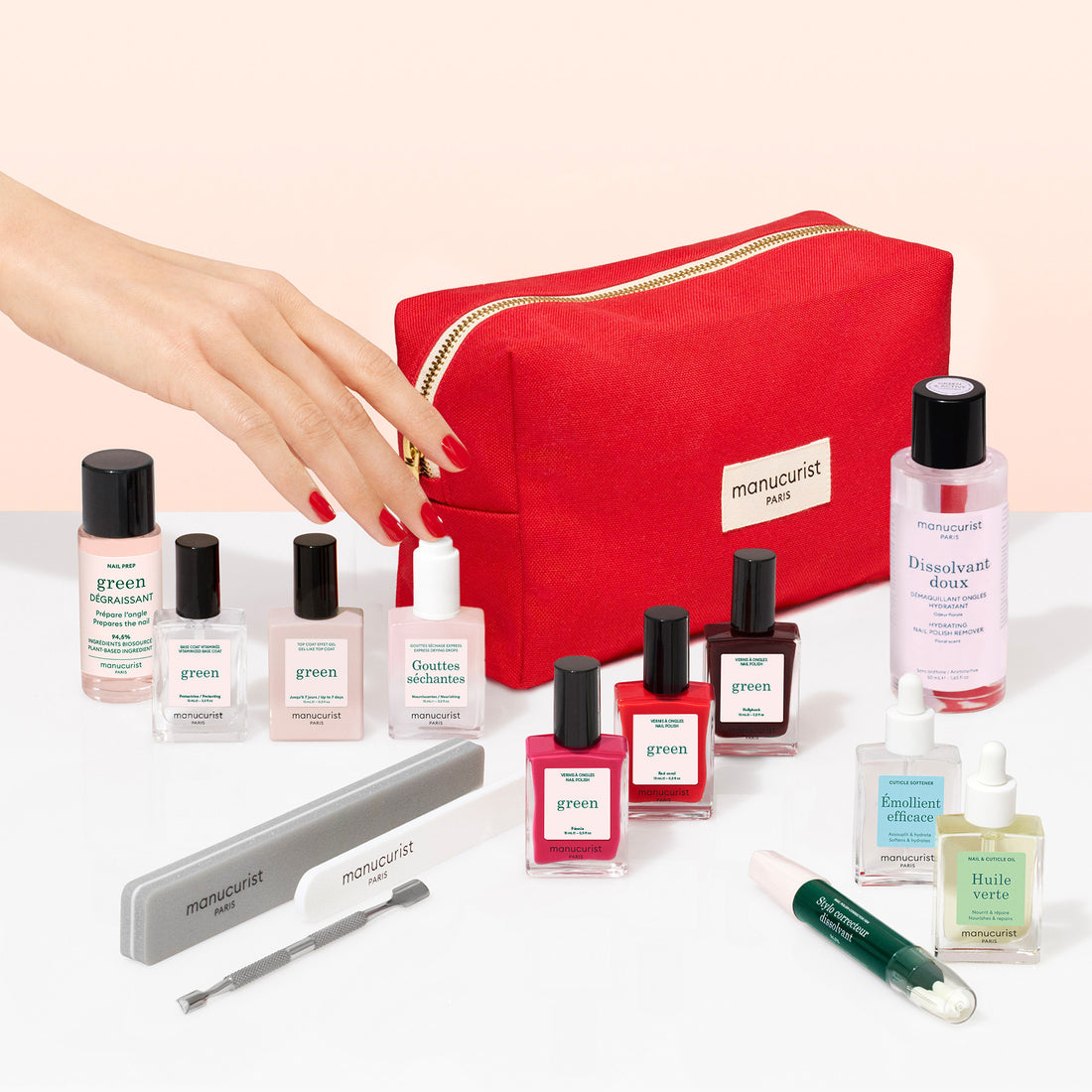 A hand with red nails holds the Manucurist Ultimate Kit—a red manicure bag arranged on a white surface alongside various nail polishes, a vitaminized base coat, nail care bottles, nail files, a cuticle tool, and a green nail pen.