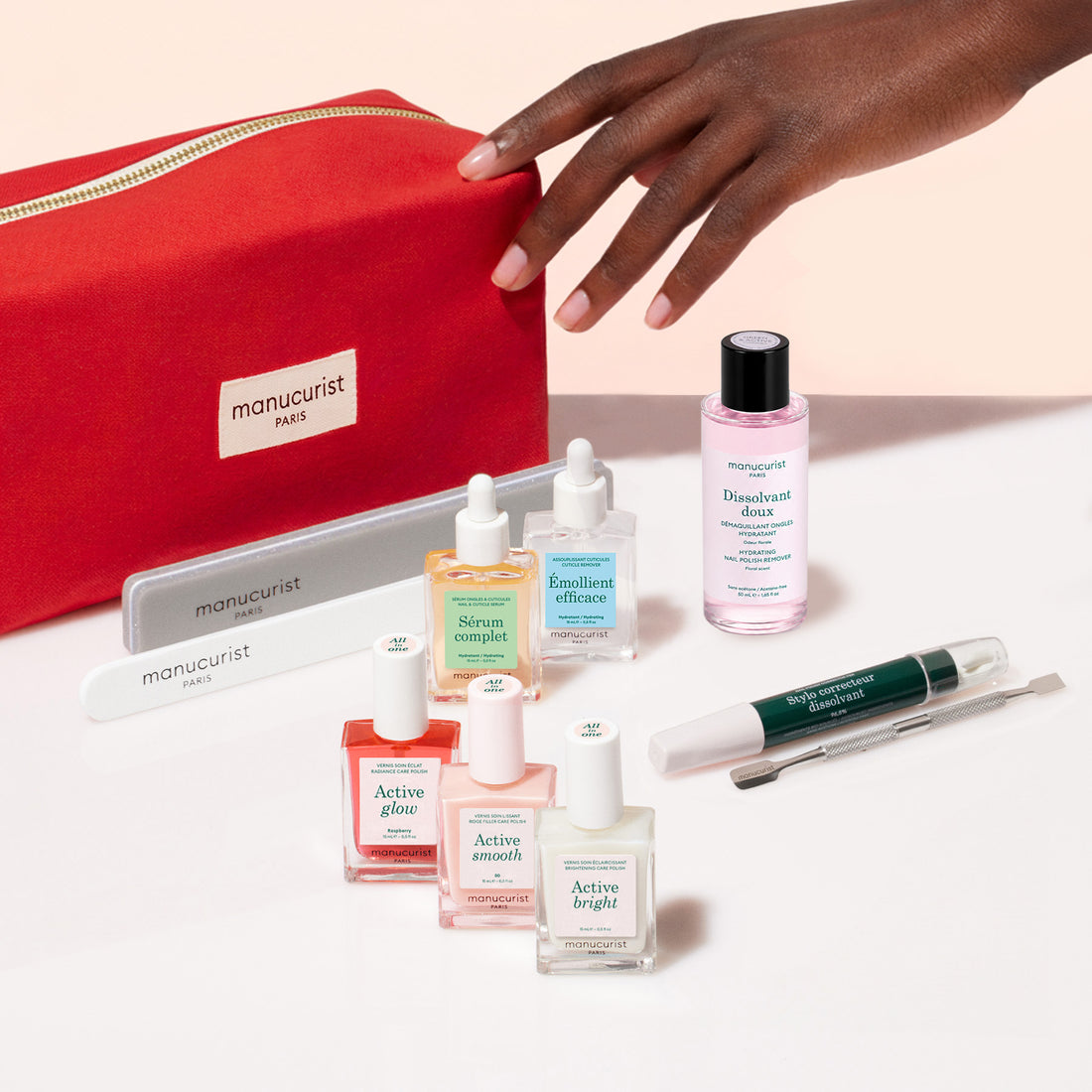 A hand reaches toward the vibrant red Ultimate Active Manicure Kit by Manucurist, surrounded by an array of nail care essentials—including nail polishes, files, a gentle cuticle remover, nourishing serums, polish remover, and professional manicure tools—all beautifully arranged on a pale surface to help you achieve healthy nails.