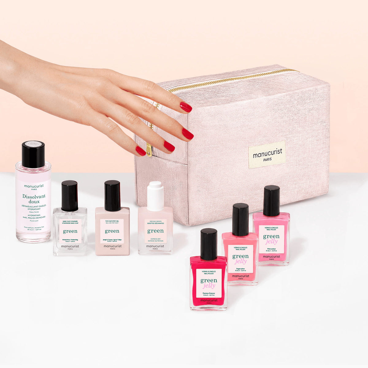 A hand with red nails reaches toward bottles of Manucurist’s Starter Kit - Jelly Nail Polish and a glittery pink manicure bag labeled Manucurist Paris. Eight plant-based nail care products are arranged neatly on a white surface.