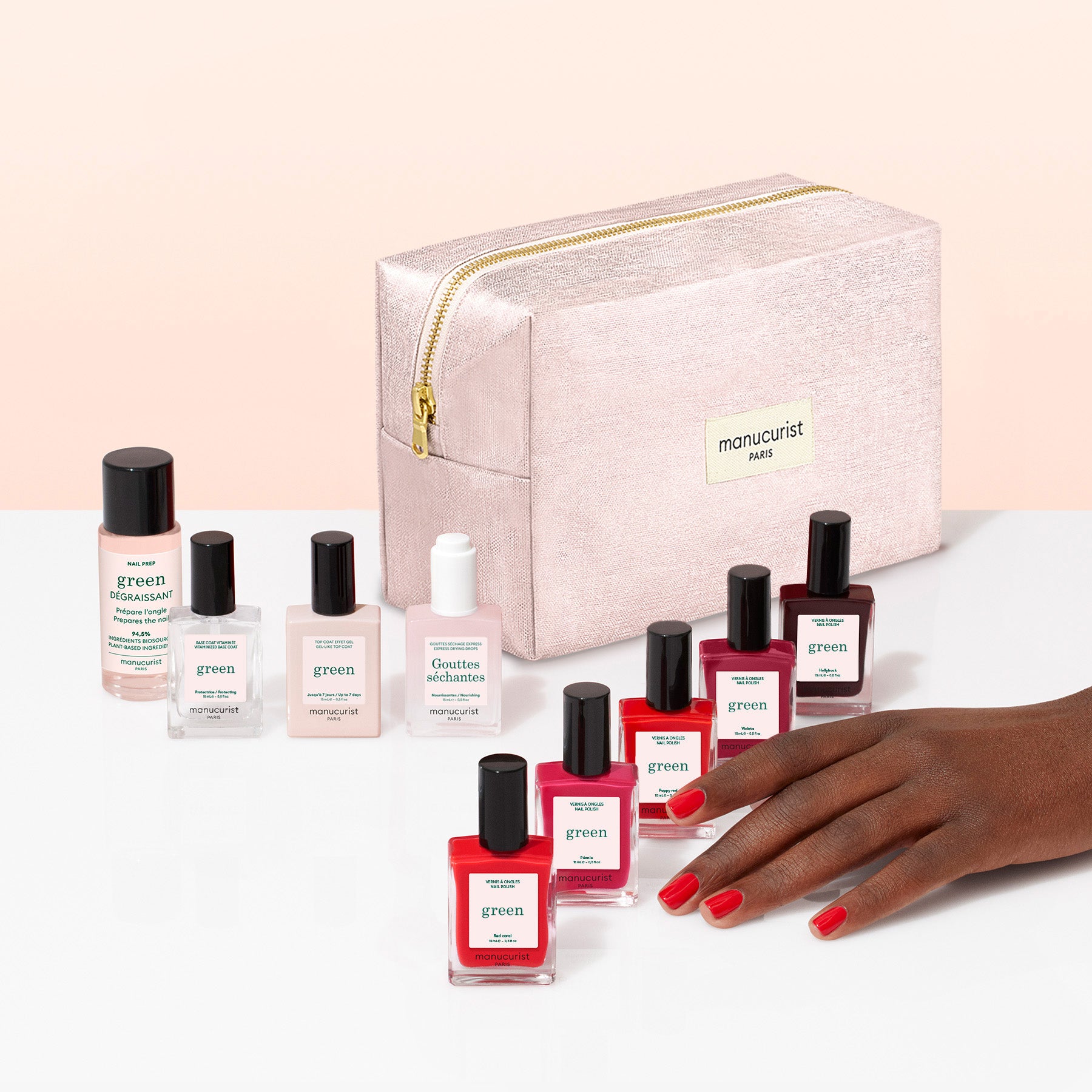 A hand with red nails—painted using a plant-based polish—rests beside eight nail polish bottles and a pink zippered pouch labeled “Manucurist PARIS” from the Manucurist Super Starter Kit, all displayed on a white surface against a pale peach background.