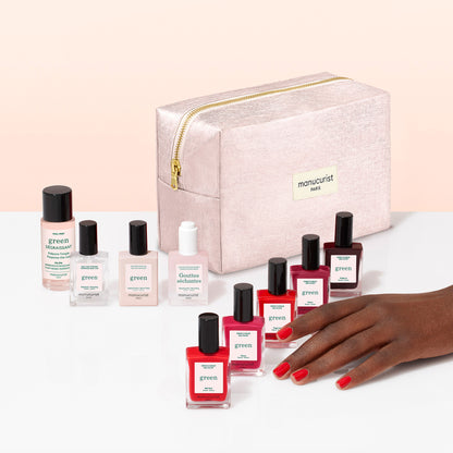 A hand with red nails—painted using a plant-based polish—rests beside eight nail polish bottles and a pink zippered pouch labeled “Manucurist PARIS” from the Manucurist Super Starter Kit, all displayed on a white surface against a pale peach background.