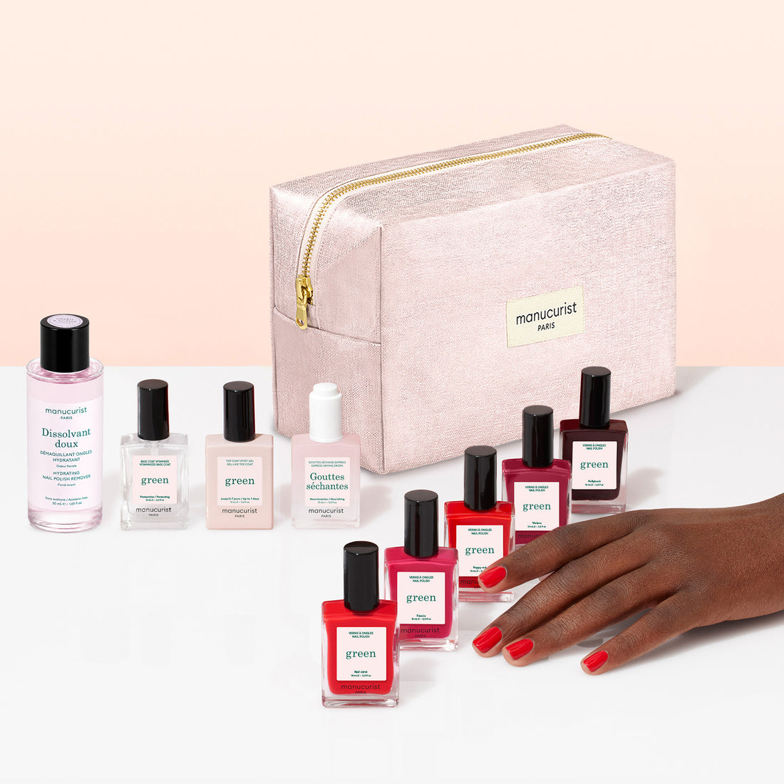 A hand with red nail polish rests beside several bottles of Manucurist plant-based nail polish in various shades, accompanying nail care products, and a light pink zippered cosmetic bag labeled manucurist Paris—all part of the Super Starter Kit—beautifully arranged on a white and pink surface, making it perfect for a Green manicure or achieving a gel effect manicure at home.