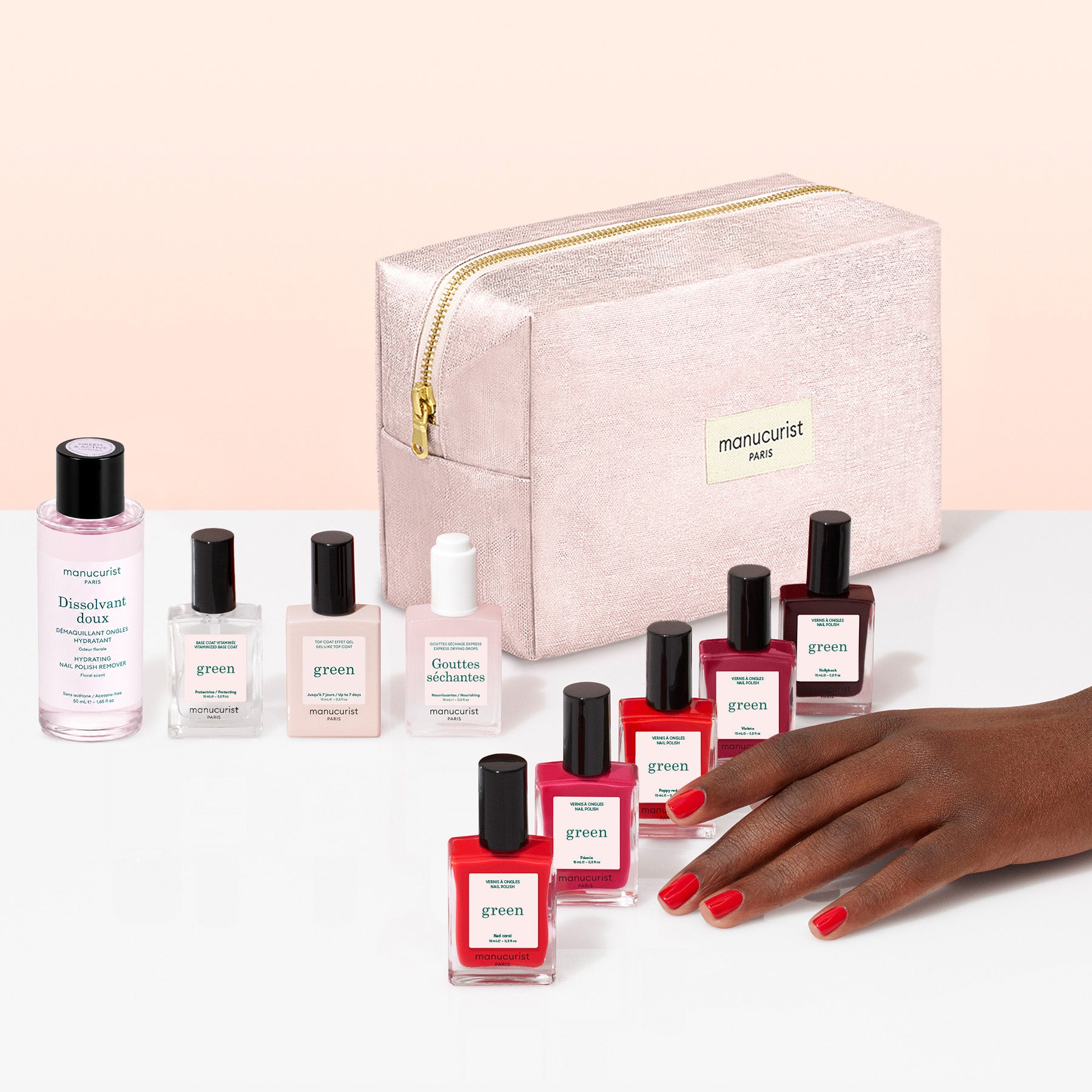 A hand with red nail polish rests beside several bottles of Manucurist plant-based nail polish in various shades, accompanying nail care products, and a light pink zippered cosmetic bag labeled manucurist Paris—all part of the Super Starter Kit—beautifully arranged on a white and pink surface, making it perfect for a Green manicure or achieving a gel effect manicure at home.