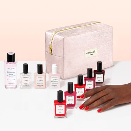 A hand with red nail polish rests beside several bottles of Manucurist plant-based nail polish in various shades, accompanying nail care products, and a light pink zippered cosmetic bag labeled manucurist Paris—all part of the Super Starter Kit—beautifully arranged on a white and pink surface, making it perfect for a Green manicure or achieving a gel effect manicure at home.