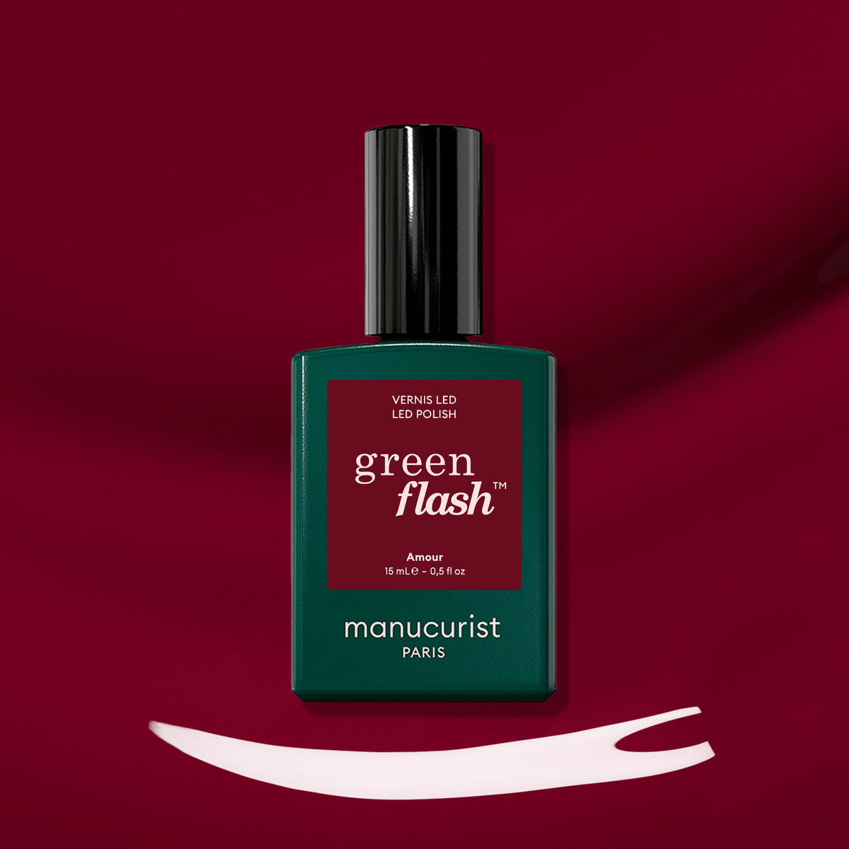 Un flacon de vernis LED Amour de Manucurist repose sur une surface rouge profond. En dessous, un trait de vernis blanc met en valeur la teinte bordeaux lumineuse de ce vernis gel emblématique.