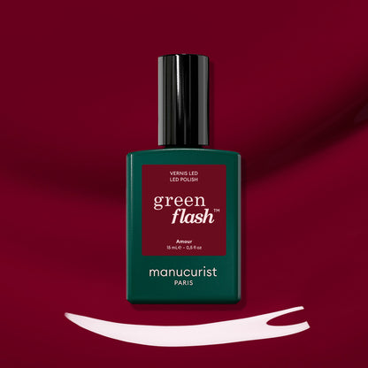 Un flacon de vernis LED Amour de Manucurist repose sur une surface rouge profond. En dessous, un trait de vernis blanc met en valeur la teinte bordeaux lumineuse de ce vernis gel emblématique.