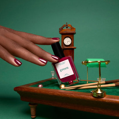 Une main aux ongles vernis d’Amour, un bordeaux lumineux, posée au-dessus d’un billard miniature à côté d’un flacon de la même teinte.