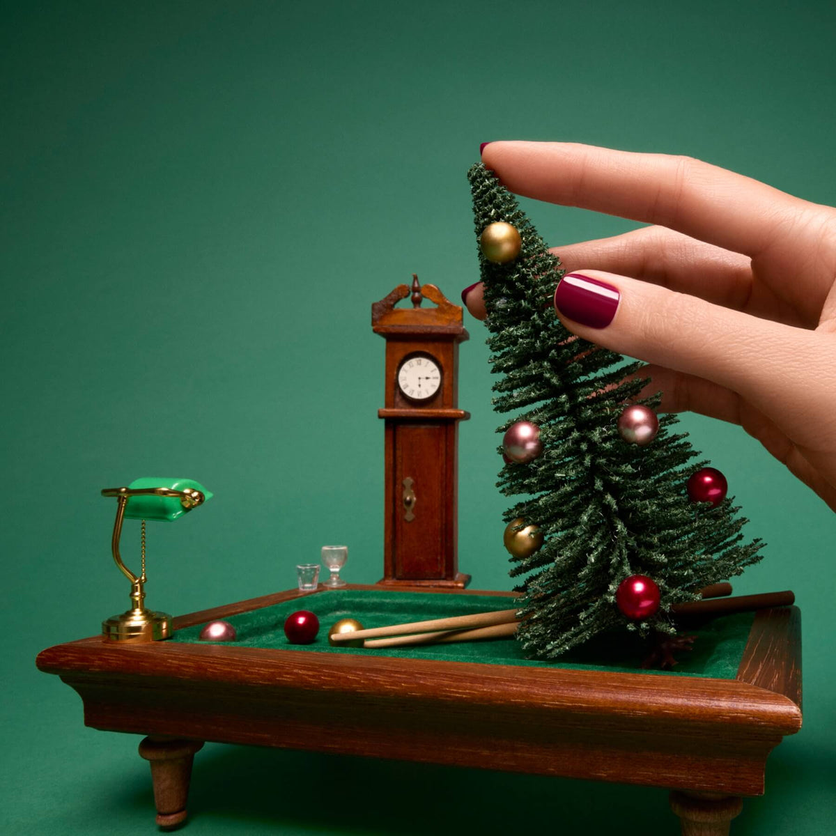 Une main aux ongles vernis d’un bordeaux lumineux avec la teinte Amour de Manucurist pose un sapin de Noël miniature sur un petit billard vert. En arrière-plan, une petite lampe, deux queues de billard et une horloge se détachent sur un décor vert inspiré de la gamme Green™.