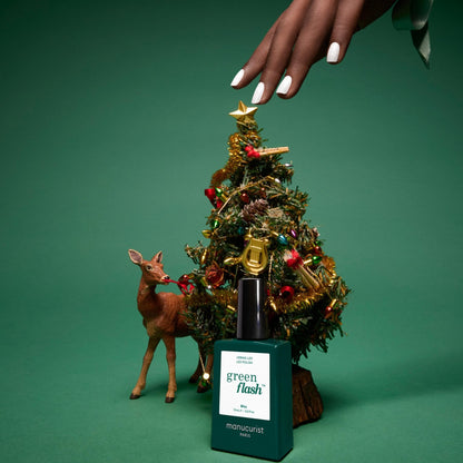 A hand with manicured nails reaches over a decorated miniature Christmas tree beside a small deer figurine. In front is a bottle of Manucurist Green Flash gel polish in the shade “Bliss” on a green background, its iridescent white hues adding an elegant festive touch to the scene.