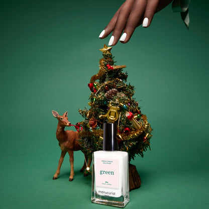 A hand with opaque pearly white nail polish reaches toward a small decorated Christmas tree beside a reindeer figurine. In front, a bottle of Manucurist Green™ nail polish in the shade Bliss stands on a green background.