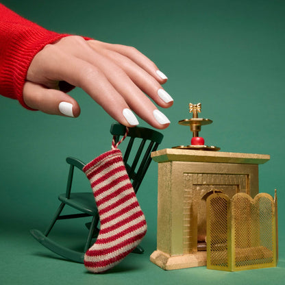 A hand with Manucurist’s Bliss iridescent white gel polish hovers over a small green rocking chair, a red and white striped stocking, and a miniature gold fireplace against a green background.