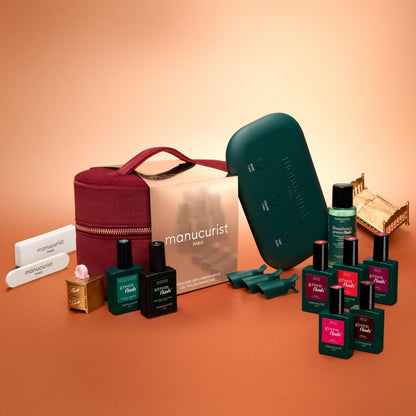 The Manucurist Green Flash™ Super Starter Kit Gift Set—including several bottles of nail polish, a burgundy carrying case, a green nail lamp, and nail files—is displayed alongside miniature gold dollhouse furniture on an orange and beige gradient background, providing everything you need for salon-style manicures and long-lasting nails at home.