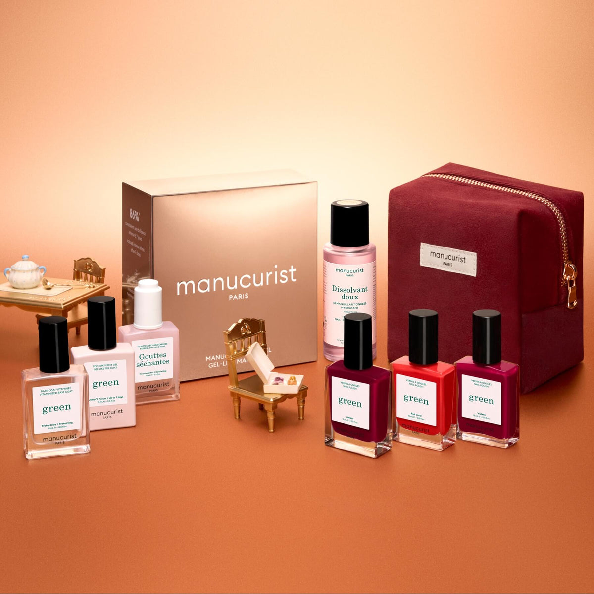 The Manucurist Green™ Starter Kit Gift Set is displayed alongside miniature furniture on a peach background, featuring naturally derived nail care essentials—including nail polish, remover, and a red velvet pouch—making it perfect for curating your own gel effect manicure or assembling a delightful holiday gift set.