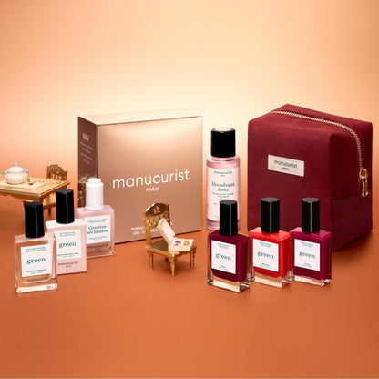 The Manucurist Green™ Starter Kit Gift Set is displayed alongside miniature furniture on a peach background, featuring naturally derived nail care essentials—including nail polish, remover, and a red velvet pouch—making it perfect for curating your own gel effect manicure or assembling a delightful holiday gift set.