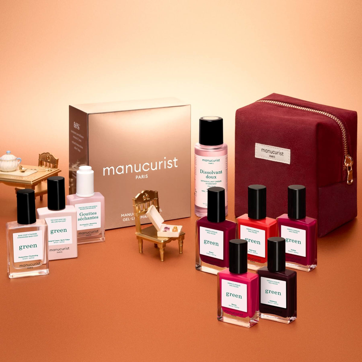 The Manucurist Green™ Super Starter Kit Gift Set—including naturally derived gel effect nail polishes, a gentle remover bottle, a gold box, and a red velvet case—is artfully arranged with miniature chairs and a table on a peach background, creating an inviting scene to enjoy your manicure or prep your nails with this luxe starter kit.