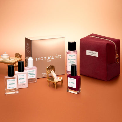 The Manucurist Green™ Discovery Kit Gift Set, featuring naturally derived nail polishes, remover, and a red pouch, is displayed with miniature furniture on a peach background. A gold Manucurist box sits in the back, making this set an ideal holiday gift.
