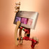 A whimsical display features the Manucurist Petite Manucurist Trio, a holiday gift manicure set with natural origin children’s nail polish in purple and pink shades, showcased alongside a toy bear, a gingerbread sled, and wooden soldier legs. The background is a warm orange gradient.