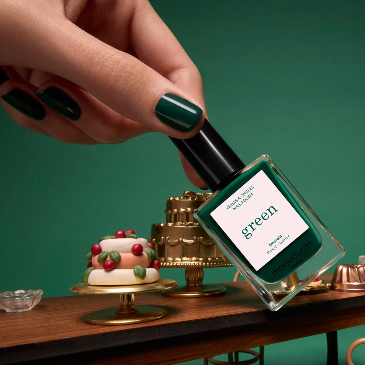 A hand with green-painted nails, adorned in Manucurist’s Emerald deep forest green polish, holds a Manucurist Green Flash LED gel polish bottle above a table with miniature cakes and desserts, all set against a rich green background.