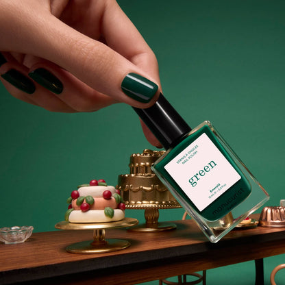 A hand with green-painted nails, adorned in Manucurist’s Emerald deep forest green polish, holds a Manucurist Green Flash LED gel polish bottle above a table with miniature cakes and desserts, all set against a rich green background.