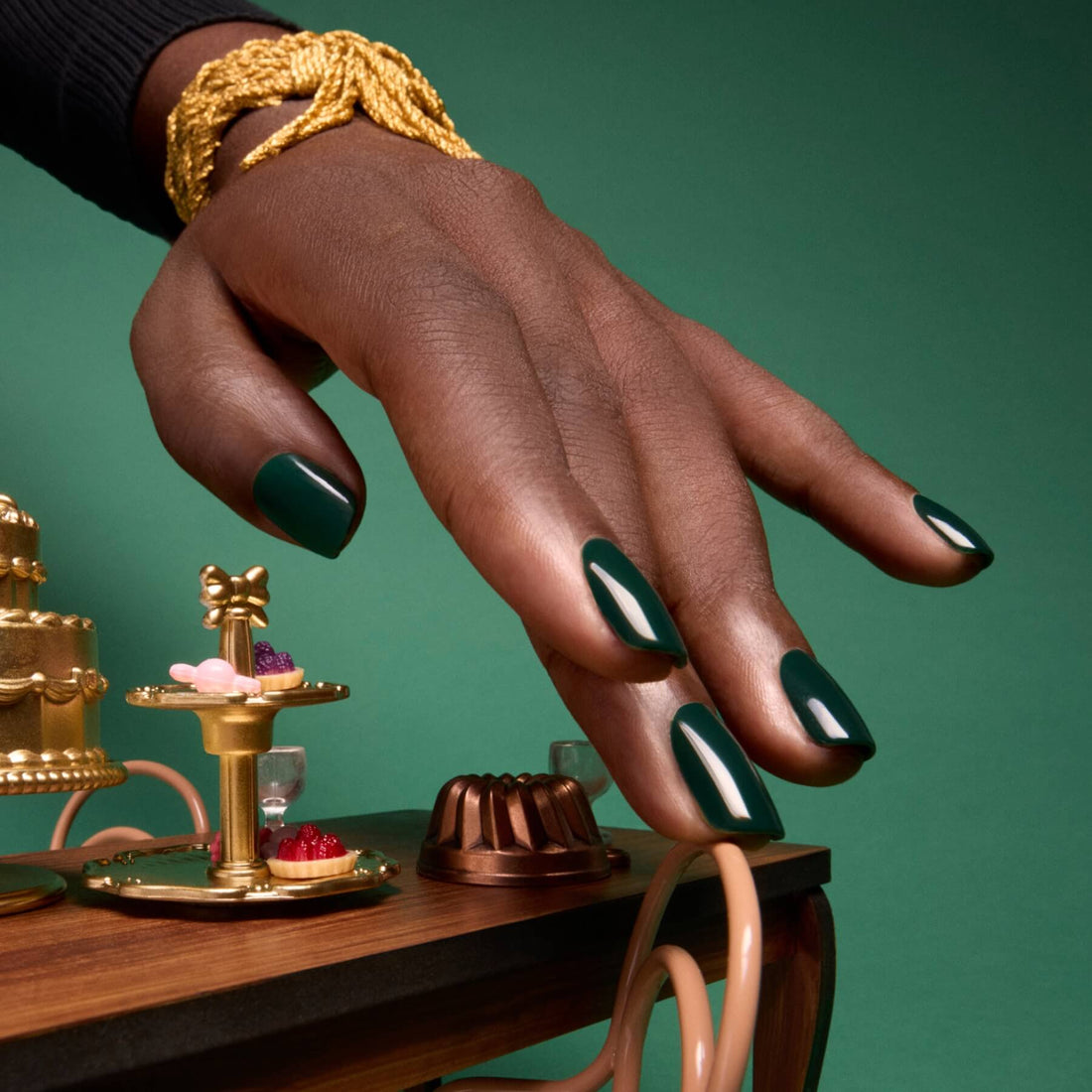 A hand adorned with Manucurist’s “Emerald” glossy, deep forest green nail polish and gold bracelets reaches toward a miniature dessert table with cakes and pastries, set against a matching green background.