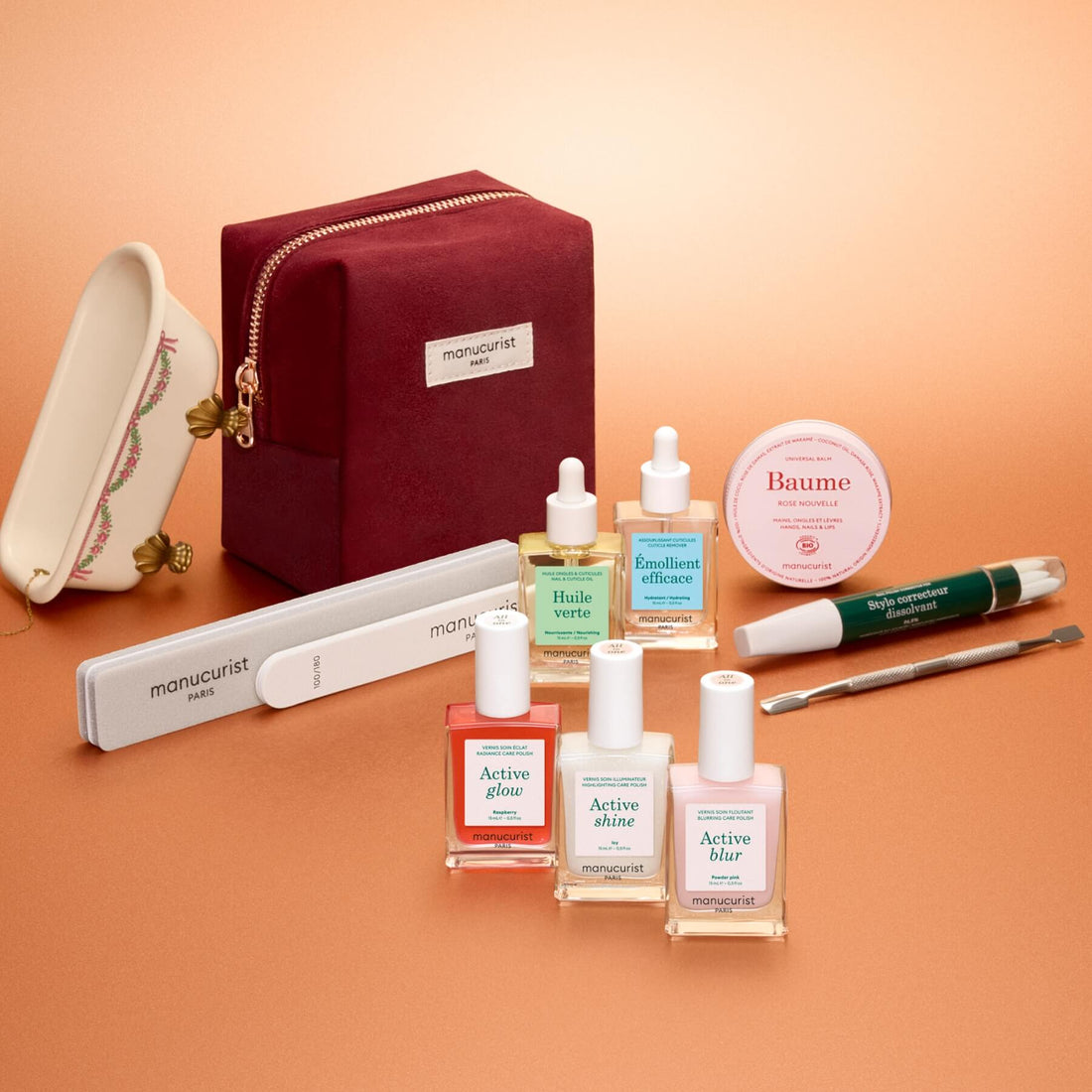 The Manucurist Active™ Ultimate Gift Set, displayed on a peach background, features nail polishes, nourishing oils, balm, a buffer, nail file, manicure tool, a petite porcelain tray, and a burgundy zip pouch adorned with the Manucurist logo—an ideal way to upgrade your manicure routine or give as a thoughtful holiday gift to any nail care enthusiast.