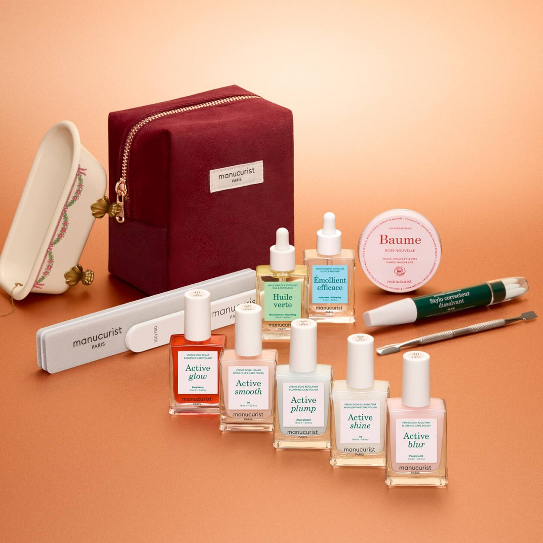 The Manucurist Active™ Super Ultimate Gift Set features a burgundy pouch, nail care tools, and eight bottles of nail and skin care products—all elegantly arranged on a peach background. The set includes oils, serums, creams, gentle nail remover, and polishes, making it a perfect holiday gift for anyone who values self-care or leads an active manicure routine.
