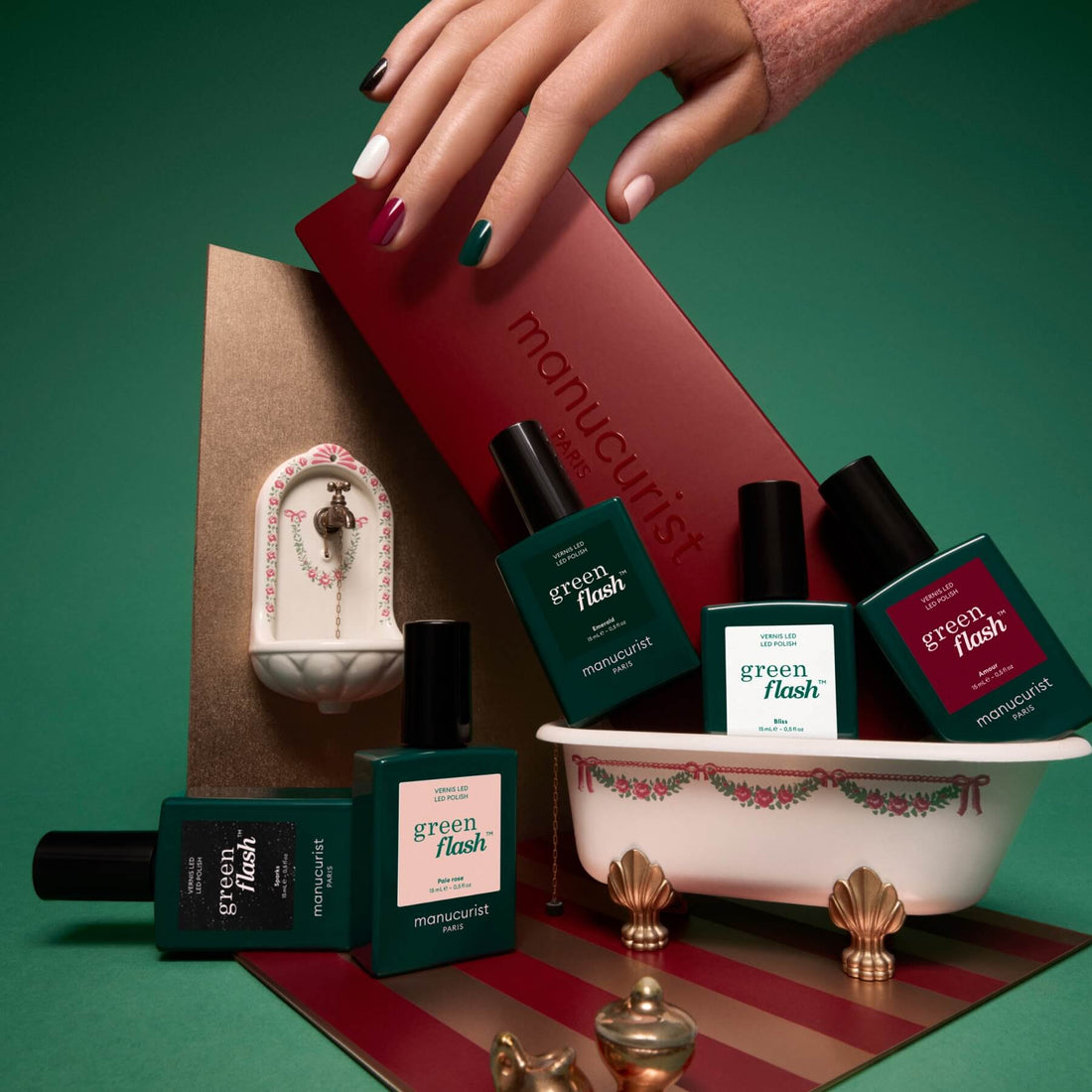 A hand with multicolored nails hovers over a gift box labeled with the Manucurist brand. Six nail polish bottles from the Curated Color Collections, inspired by Green Flash color palettes, are arranged in and around a small decorative tub while a tiny ornate sink sits in the background. The green and red color scheme highlights the latest manicure trends.