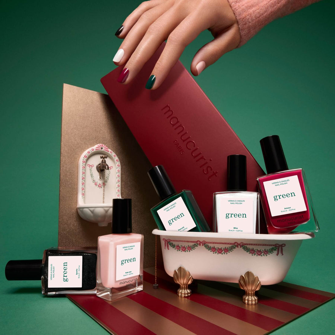 A hand with multicolored nails, featuring the latest nail trends and vibrant shades from Manucurist’s Curated Color Collections, holds a red box above mini bottles displayed in a decorative bathtub, set against a pink and gold backdrop.