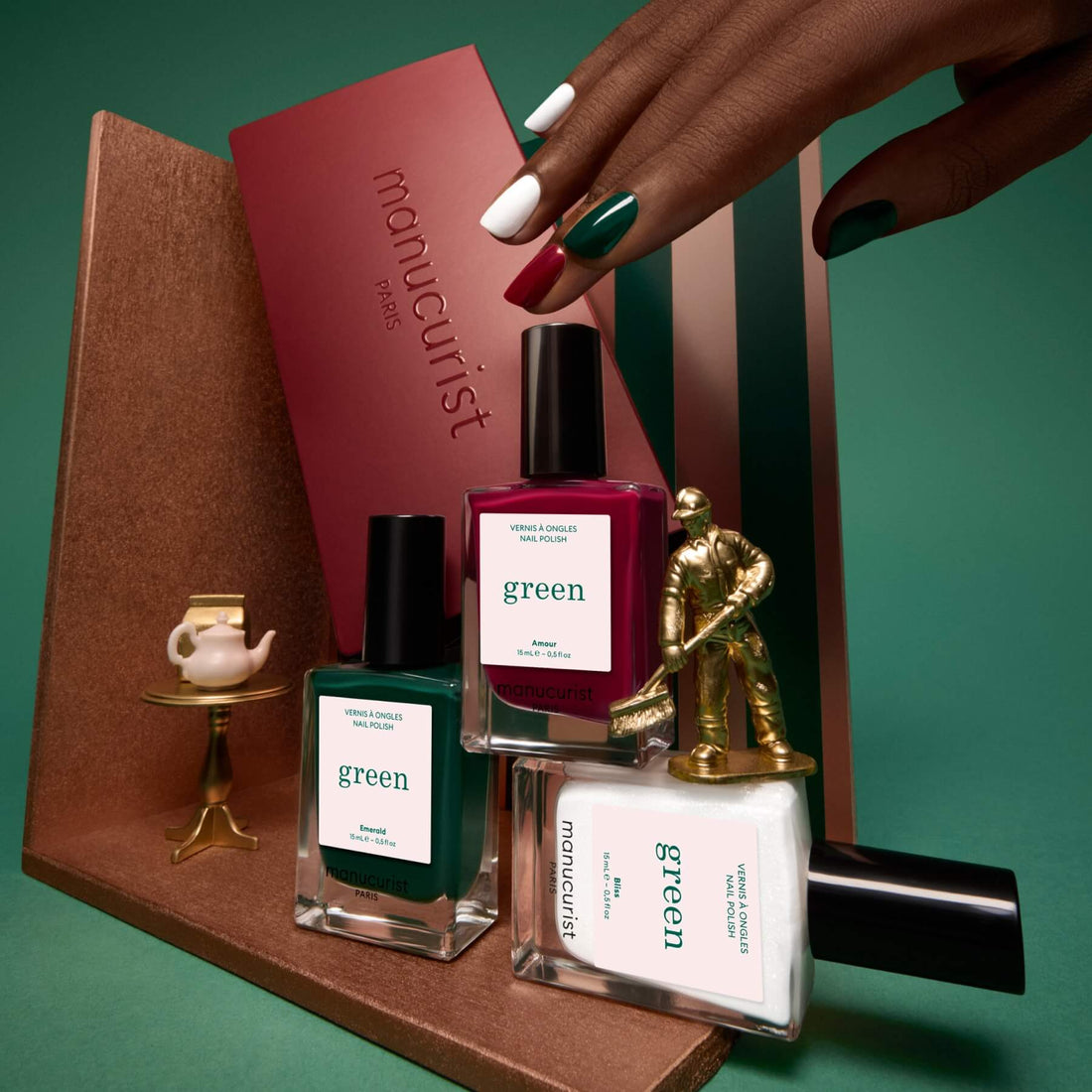 A hand with painted nails holds a maroon box labeled “Curated Trios” by Manucurist, displayed beside three Manucurist nail polish bottles, a miniature gold golfer, and a tiny tea set on a gold stand—all artfully arranged on geometric platforms to highlight this elegant collection of manicure trend-inspired color palettes.
