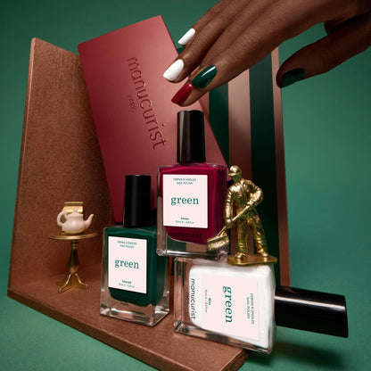 A hand with painted nails holds a maroon box labeled “Curated Trios” by Manucurist, displayed beside three Manucurist nail polish bottles, a miniature gold golfer, and a tiny tea set on a gold stand—all artfully arranged on geometric platforms to highlight this elegant collection of manicure trend-inspired color palettes.
