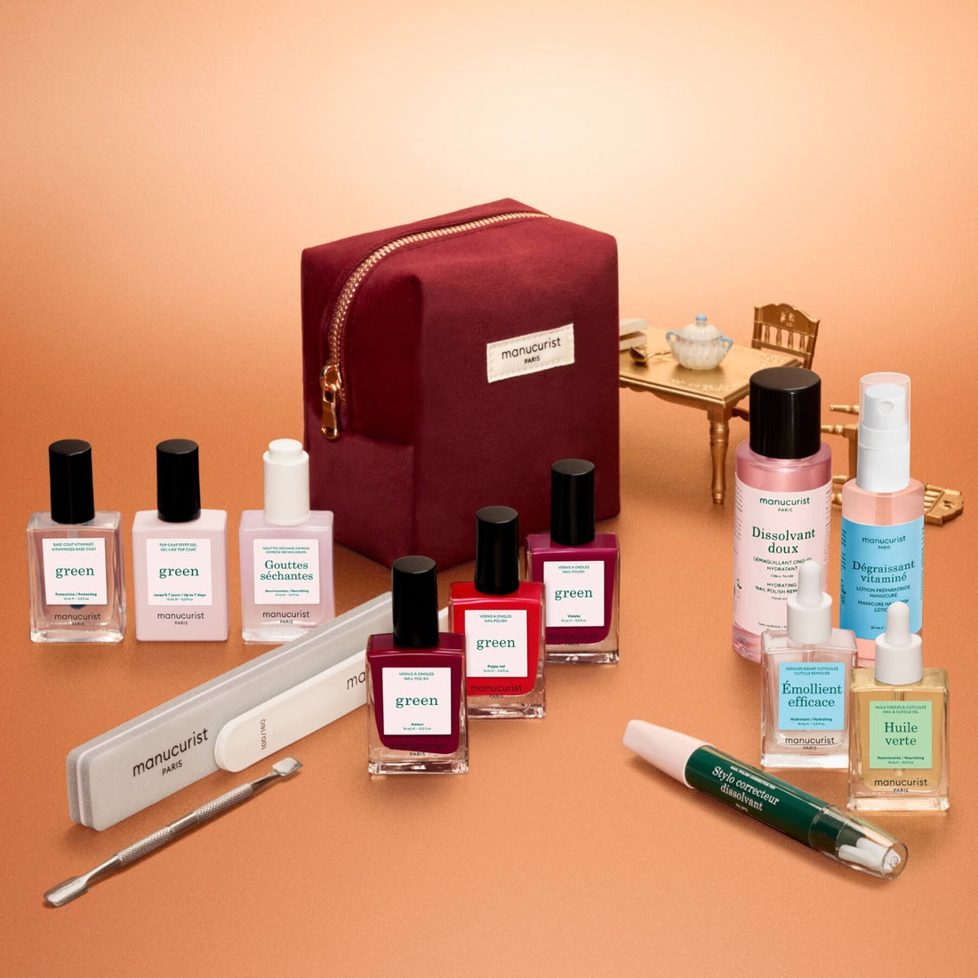 Le coffret kit complet Green™ de Manucurist est présenté avec élégance : une trousse de manucure bordeaux entourée de vernis effet gel, de vernis d’origine naturelle, de soins, de limes et d’accessoires de manucure professionnels. À l’arrière-plan, une table et une chaise dorées miniatures, accompagnées d’une théière et d’une tasse, apportent du charme à la scène. Tous les produits sont soigneusement disposés pour une séance de manucure à la maison.