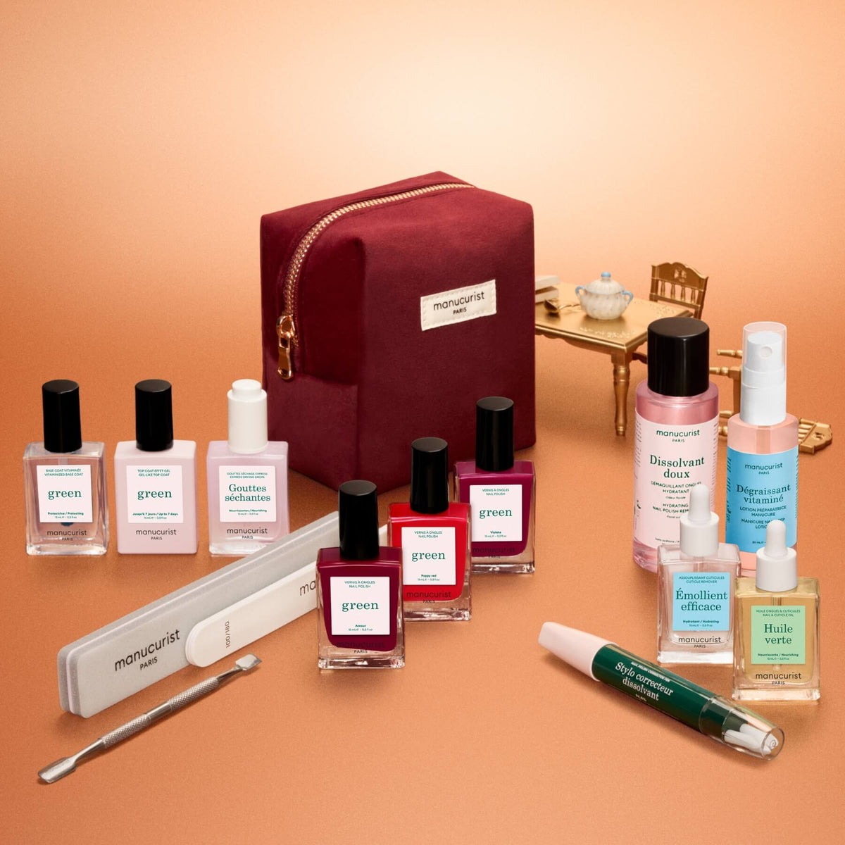 The Manucurist Green™ Ultimate Kit Gift Set is elegantly displayed, featuring a burgundy manicure bag surrounded by gel effect polish, naturally derived nail polishes, treatments, nail files, and professional manicure tools. In the background, a miniature golden table and chair with a teapot and cup add charm to the scene. All products are thoughtfully arranged—ideal for creating an at-home manicure experience.