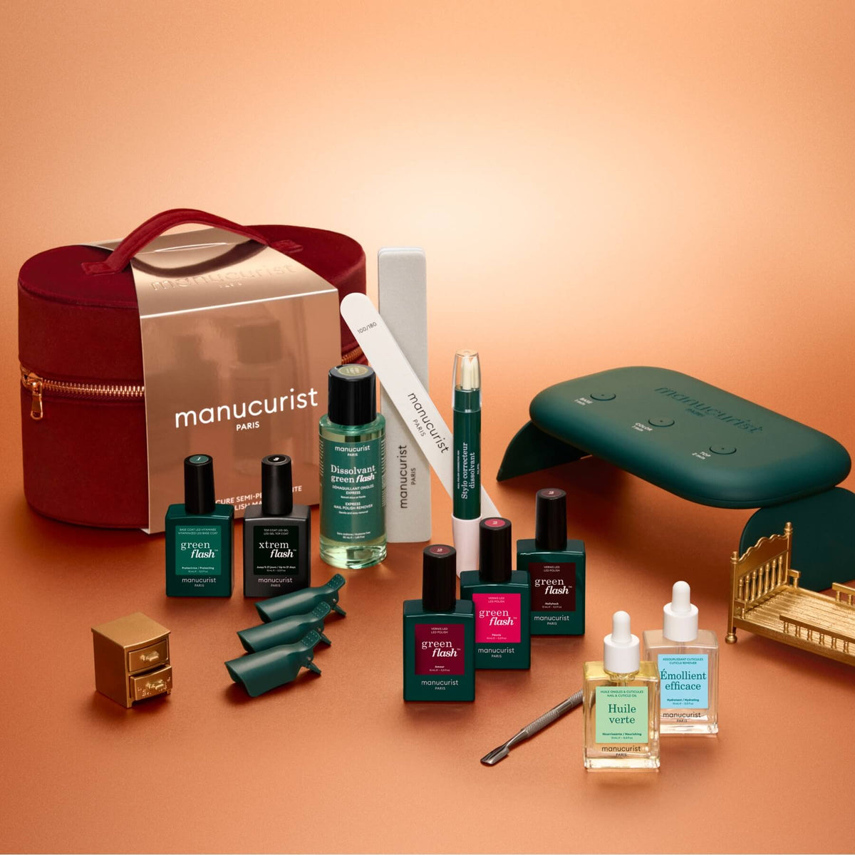 Achieve a salon-quality manicure at home with the Manucurist Green Flash™ Ultimate Kit Gift Set, which includes premium nail polishes, remover, filing tools, essential nail accessories, mini bottles, a stylish dark green LED nail lamp for perfect results, and an elegant red carrying case—all beautifully arranged on a bronze-toned surface.