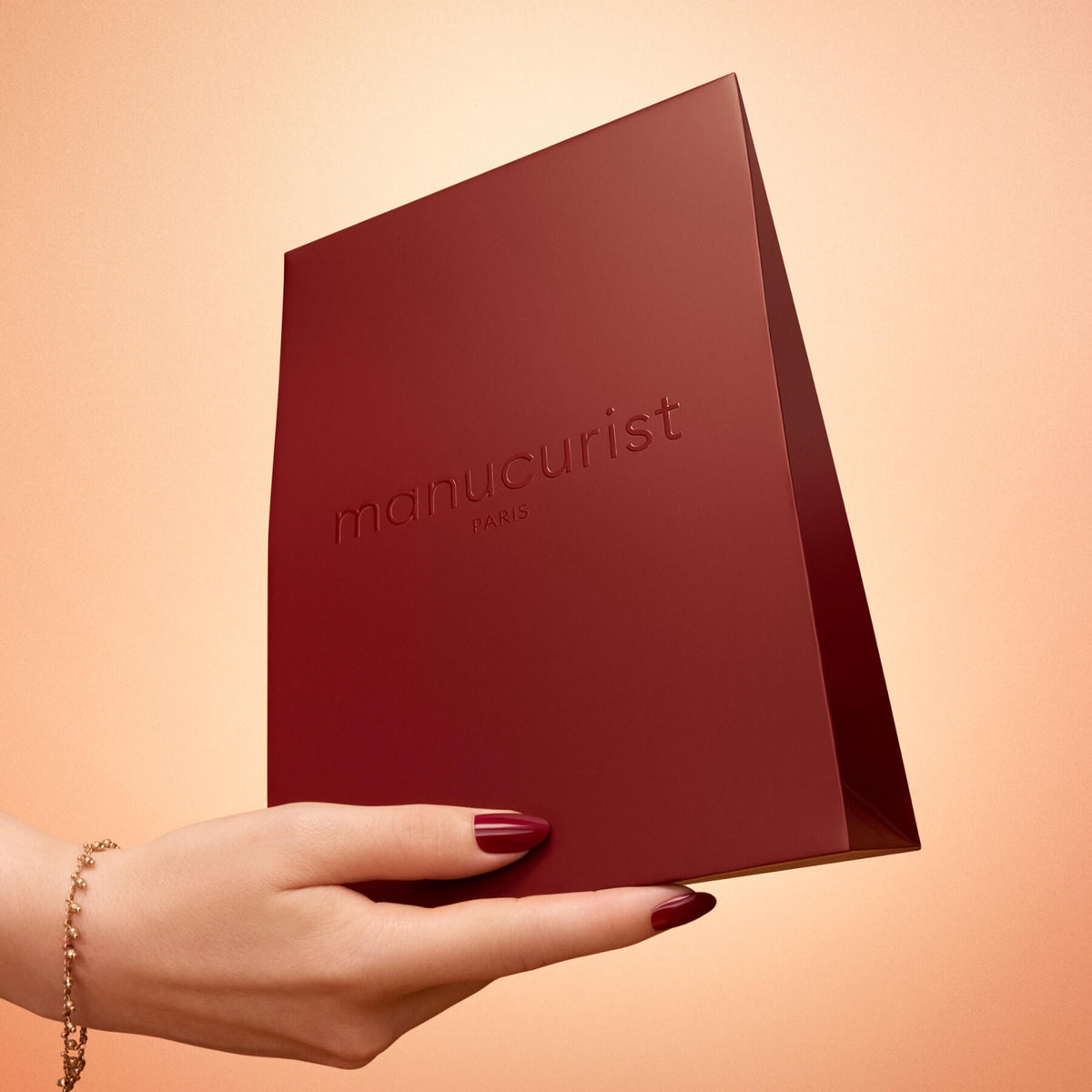 A hand with red-painted nails and a gold bracelet holds the Small Holiday Gift Bag by Manucurist, a matte burgundy bag embossed with “manucurist PARIS,” set against a peach background.